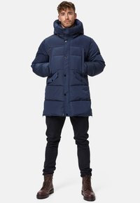 Doudoune marine avec un col haut, deux poches avant, des fermetures à bouton et un motif matelassé. Portée avec un pantalon noir et des bottes marron.