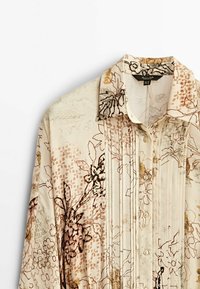 Blouse à manches longues beige avec plissé à l'avant et motif floral en tons marron, noir et moutarde, dotée d'un col pointu et d'une fermeture à boutons.