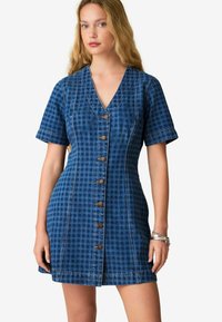 Abito in denim con motivo a quadri, scollo a V, maniche corte e bottoni marroni davanti. Colore blu con una finitura testurizzata.