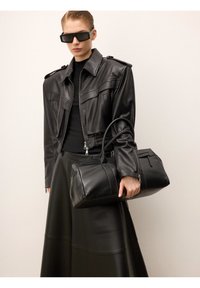 Fekete bőrkabátot és szoknyát viselő nő, fekete bőr sporttáskával a kezében, nagy téglalap alakú napszemüveget viselve.