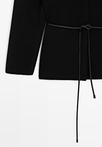 Pull en maille noir à manches longues avec une fine ceinture noire nouée en nœud à la taille, tombant droit vers le bas.