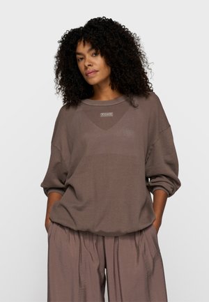 Kobieta z kręconymi włosami, ubrana w luźną, teksturowaną brązową bluzkę z długim rękawem oraz pasujące szerokie spodnie, mająca ręce w kieszeniach.