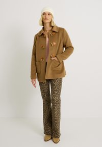 Abrigo de lana marrón con acentos de cuero, diseño de doble botonadura, grandes bolsillos, combinado con pantalones de campana de estampado de leopardo y tacones marrones.