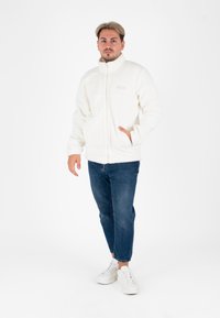 Witte fleecejas met hoge kraag, ritsluiting en zijzakken, gecombineerd met blauwe jeans en witte sneakers. De textuur is zacht en vederachtig.