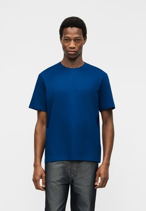Man staat met het gezicht naar voren, draagt een effen blauw t-shirt met korte mouwen en donkergrijze broek tegen een witte achtergrond.