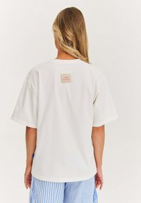 Bílá bavlněná tričko s krátkým rukávem, na zadní straně má čtvercovou etiketu. Texturovaná tkanina, volný střih, v kombinaci s pruhovanými modrými šortkami.