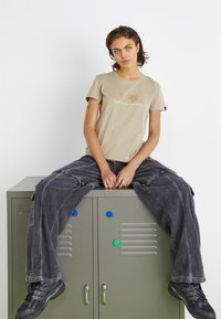 béžové bavlnené tričko s textom a logom "ALPHA INDUSTRIES", v kombinácii s voľnými tmavošedými cargo nohavicami a čiernymi teniskami, sediace na šedom zámku.