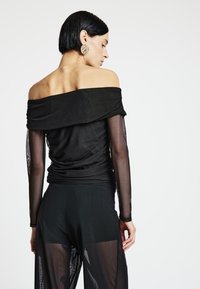 Schwarzes schulterfreies Oberteil mit strukturierter, drapierter Stoffführung und transparenten, langen Ärmeln, kombiniert mit schwarzen, fließenden, transparenten Hosen.