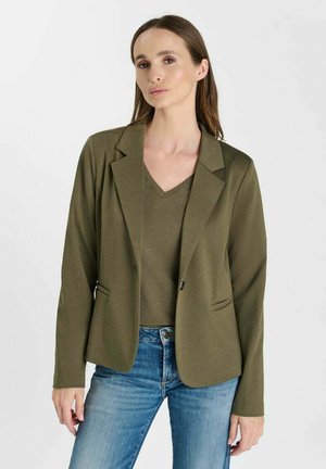 Frau mit langen braunen Haaren, die einen olivgrünen Blazer, ein passendes V-Ausschnitt-Top und blaue Jeans vor einem schlichten hellen Hintergrund trägt.