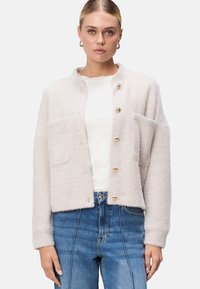 Lysbeige tekstureret cardigan med rund krave, guldknapper og to forlommer, parret med en hvid skjorte og blå jeans.