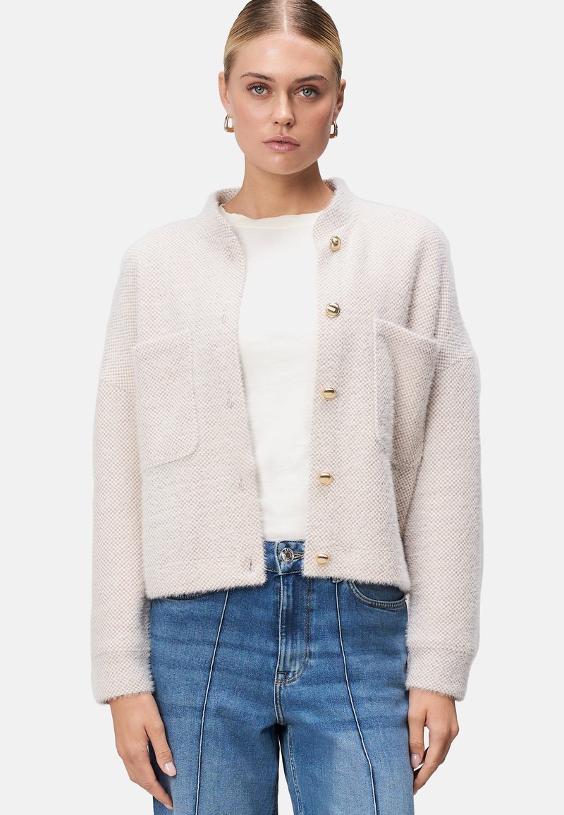Lysbeige tekstureret cardigan med rund krave, guldknapper og to forlommer, parret med en hvid skjorte og blå jeans.