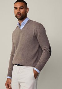 Ruskea V-pääntie neule pehmeästä kankaasta, yhdistettynä vaaleansiniseen raidalliseen kauluspaitaan. Ominaisuuksiin kuuluu muotoiltu hiha ja klassinen muotoilu.