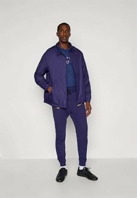 Lilla kerge jakk, millel on nööbid ees, koos tume sinise T-särgiga ja sobivate lilla dressipükstega, moodustades harmoonilise spordiliku välimuse.