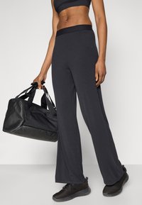 Čierne široké tréningové nohavice s textúrovaným pásom, doplnené o čiernu športovú tašku a čierne sieťované tenisky, držené v ruke.