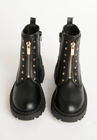 Botas negras hasta el tobillo con cremalleras laterales, detalles de tachuelas, cuero texturizado y suelas de goma gruesas. Presenta un lazo para facilitar su uso.