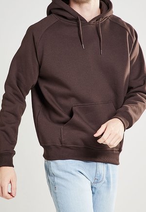 Mann trägt einen dunkelbraunen Hoodie mit Fronttasche und Kordelzug, kombiniert mit hellblauen Jeans, steht vor einem einfarbigen Hintergrund.