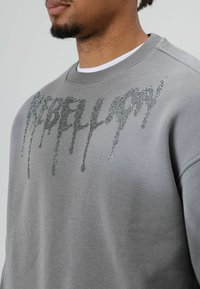 Sweat-shirt gris à texture douce, arborant un motif "REBELLION" orné de strass qui dégouline sur le devant.