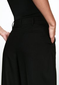 Pantalon noir à jambes larges avec une texture lisse, doté d'une poche arrière discrète et d'une ceinture ajustée. Design minimaliste et couleur classique.
