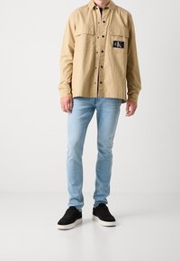 Hombre vestido con una camisa de botones beige con el logo de Calvin Klein, jeans ajustados azul claro y zapatillas negras con suelas blancas.