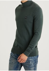 Grüner, gestrickter Rollkragenpullover mit gerippten Schulterdetails und einem kleinen schwarzen Logopatch, kombiniert mit blauen, verwaschenen Jeans.
