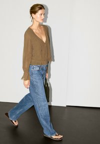 Olivenfarbene, durchsichtige Bluse mit V-Ausschnitt und Rüschenfäden, kombiniert mit hellblauen, weiten Jeans und braunen Flip-Flops, die auf einer dunklen Oberfläche gehen.
