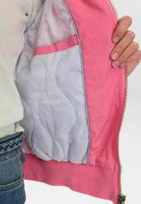 Roza zip-up jakna z notranjostjo, ki vsebuje prešito belo podlogo z majhnimi pikami, roza žepom za poudarek in rebrastimi ročki v roza barvi.