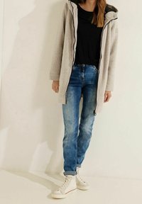 Chaqueta de forro polar gris claro con capucha, camiseta negra y vaqueros azules. Zapatillas blancas altas con detalles en rojo. Textura suave y ajuste holgado.