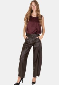 Donna in piedi con le mani nelle tasche, indossa una canotta bordeaux senza maniche, pantaloni in pelle marroni a vita alta e tacchi alti neri su sfondo bianco.