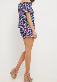 Romper blu navy a fiori con spalle scoperte, maniche corte e dettagli arricciati. Presenta un corpetto aderente e pantaloni corti morbidi, abbinati a tacchi nude.