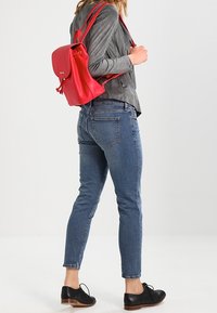 Mochila de cuero rojo, chaqueta texturizada gris, jeans ajustados de mezclilla azul y zapatos negros con cordones. La mochila cuenta con un cierre superior y tirantes ajustables.