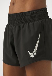 Atleta vistiendo pantalones cortos negros de Nike con un logo de swoosh blanco y estampado en el muslo izquierdo, mostrando la parte media del torso y la parte superior de las piernas.