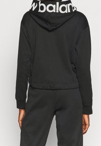 Czarna bluza z kapturem o skróconym fasonie, z prążkowanymi mankietami i białą taśmą z logo przy kołnierzu. Miękka, gładka tekstura materiału.