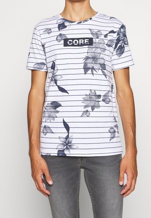Mand iført hvid kortærmet T-shirt med tynde sorte striber, blomsterprint og teksten "CORE", kombineret med grå jeans.