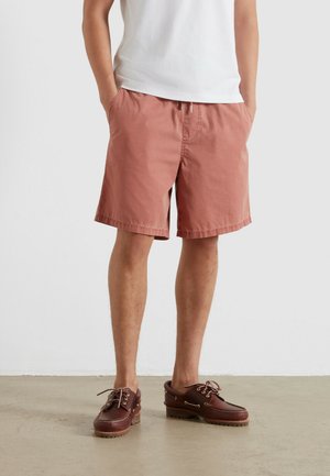 Mężczyzna ubrany w różowe szorty, białą koszulkę i brązowe skórzane buty typu boat shoes stoi na jasnej podłodze na tle białej ściany.