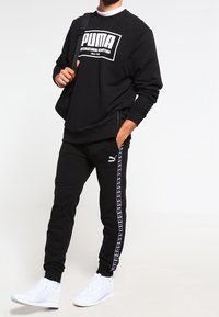 Felpa nera con logo PUMA bianco, abbinata a pantaloni della tuta neri con una banda con il marchio PUMA. Scarpe sportive bianche completano il look.