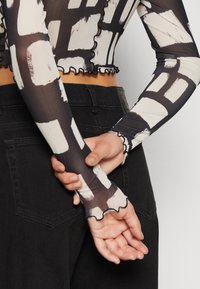 Svart genomskinlig långärmad topp med abstrakta vita mönster, med volangkanter vid ärmar och nederkant. Bärs med svarta högmidjade jeans.