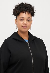 Svart hettejakke med glidelås, laget av et glatt stoff. Har sølvglidelås og en lyseblå innsiden av halsåpningen.