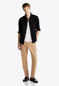 Chemise noire boutonnée sur un t-shirt blanc, pantalon beige et mocassins noirs. Le modèle se tient face à l'avant avec des cheveux courts.