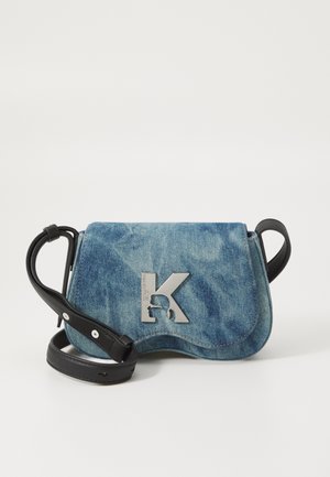 Mała torebka z niebieską klapką z denimu, srebrnym logo "K" i regulowanym czarnym skórzanym paskiem na jednolitym białym tle.