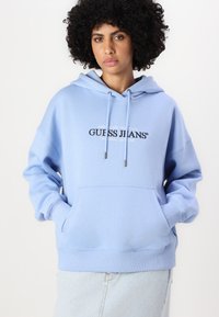 Svetlomodrá mikina s kapucňou a predným vreckom, šnúrkou na kapuci a čiernym logom "GUESS JEANS". Zmes bavlny, voľný strih a rebrované manžety.