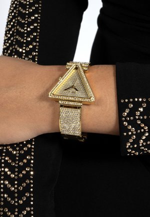 Montre dorée en forme de triangle avec des ornements en cristal portée au poignet gauche, associée à une manche noire ornée de clous dorés.