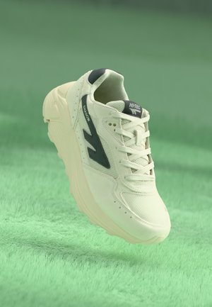 Beige treningssko med et strukturert mesh-overdel, en grønn aksentlogo, tykk hvit såle og seks hull for snøring.