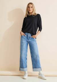 Schwarzes Strickoberteil mit Dreiviertelärmeln, kombiniert mit hellblauen, weit geschnittenen Jeans mit abgeschnittenem Saum und umgeschlagenen Bündchen, vor einem neutralen Hintergrund.