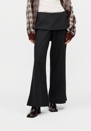 Žena nosi čierne nohavice s širokými nohavicami a asymetrickým pásovým panelom, v kombinácii s bielym topom, vzorovaným kardigánom a čiernymi členkovými čižmami na opätku.