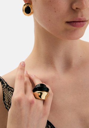 Sort og guld udtryksfuld ring med lagdelt cirkulært design. Guld øreringe med sorte detaljer. Model iført en blonder top.