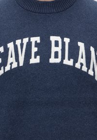 Син пуловер с големи бели букви, изписващи "LEAVE BLAN" във извита форма върху областта на гърдите.