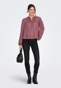 Γυναίκα όρθια, φοράει μπλούζα μακρυμάνικη σε απόχρωση μοβ, μαύρο στενό τζιν, μαύρα μποτάκια μέχρι τον αστράγαλο και κρατάει μια μαύρη τσάντα χειρός.