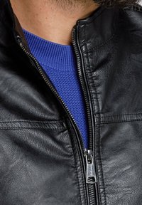 Giubbotto in pelle nera con colletto alto, dettagli con zip e finitura testurizzata, indossato sopra un maglione blu a coste.