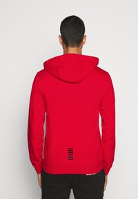 Sudadera con capucha roja y mangas largas. Cuenta con un bolsillo tipo canguro y detalles de cortes horizontales en la parte inferior de la espalda. Textura de tela suave.