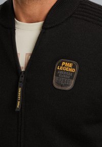 Czarna dzianinowa kurtka zapinana na zamek z ribbowanym kołnierzem, z okrągłą tkana łatką oznaczoną "PME LEGEND" w kolorze żółtym i pomarańczowym.